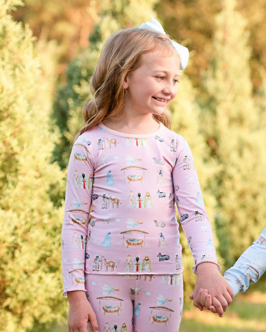 A young girl wearing a light pink long-sleeve pajama set with a colorful nativity scene print, holding hands with another child outdoors in a sunny garden setting.