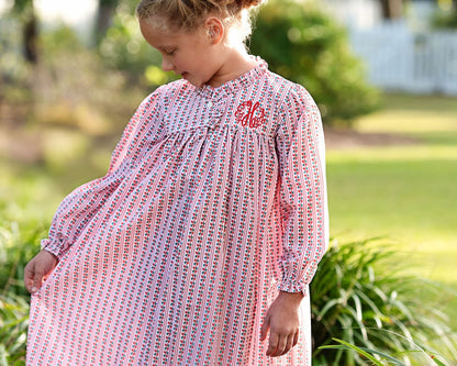 A young girl wearing a long-sleeve babydoll nightgown with a pink, white, and red striped pattern and a red monogram on the chest, standing outdoors in a sunny garden setting.