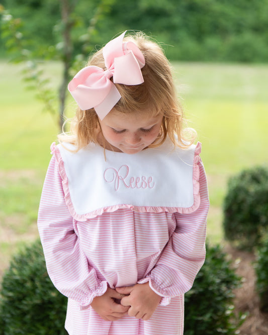 A young girl wearing a pink and white striped dress with a white ruffled bib personalized with the name 'Reese' in pink script, and a large pink bow in her hair, standing outdoors in a grassy yard.