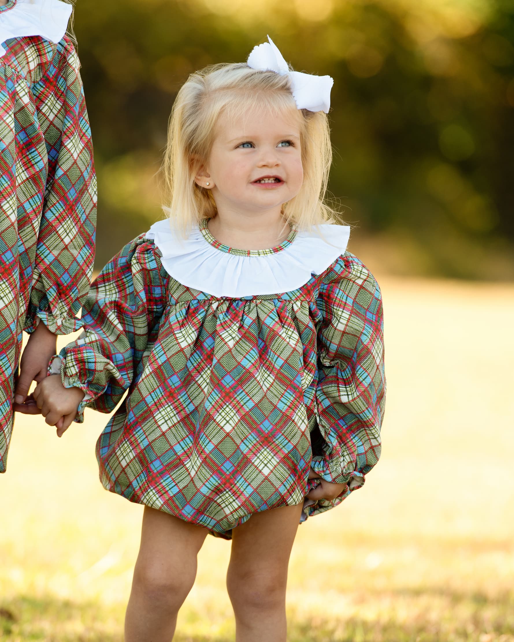A young girl wearing a plaid bubble romper with a white ruffled collar and a white hair bow, standing outdoors in a grassy field.