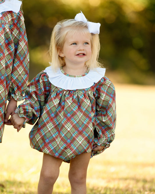 A young girl wearing a plaid bubble romper with a white ruffled collar and a white hair bow, standing outdoors in a grassy field.