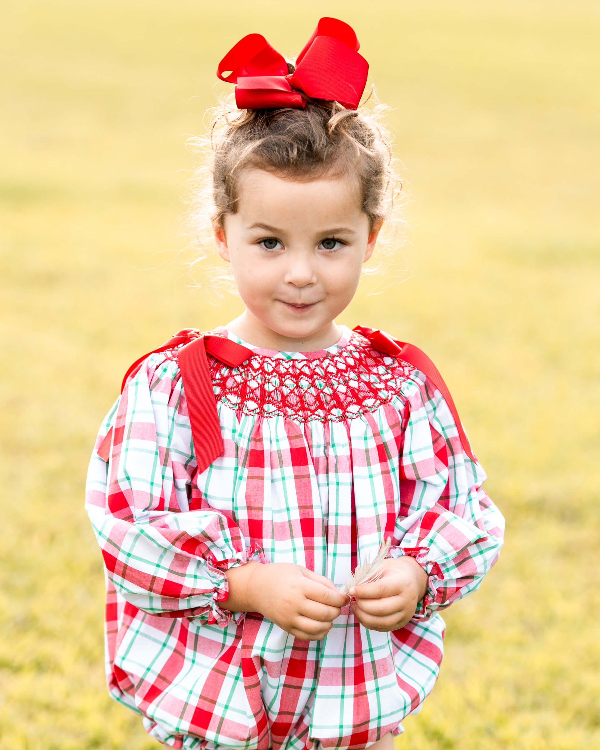 A young girl wearing a red, white, and green plaid smocked dress with red shoulder straps and a large red bow in her hair, standing in a grassy field.