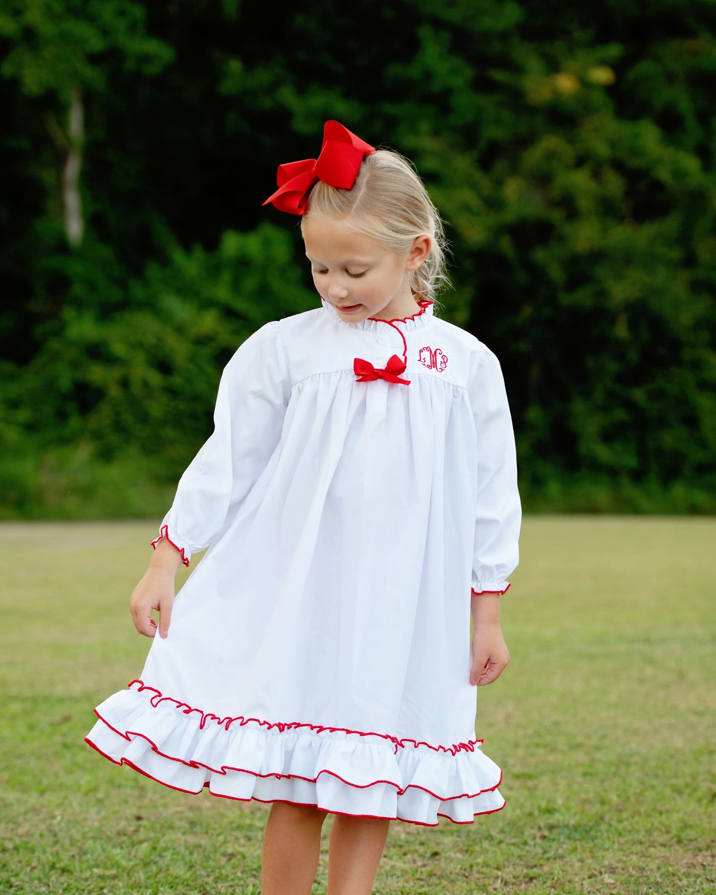 A young girl wearing a white long-sleeve dress with red trim, a red bow at the collar, and a red monogram on the chest, standing outdoors on a grassy lawn with trees in the background.