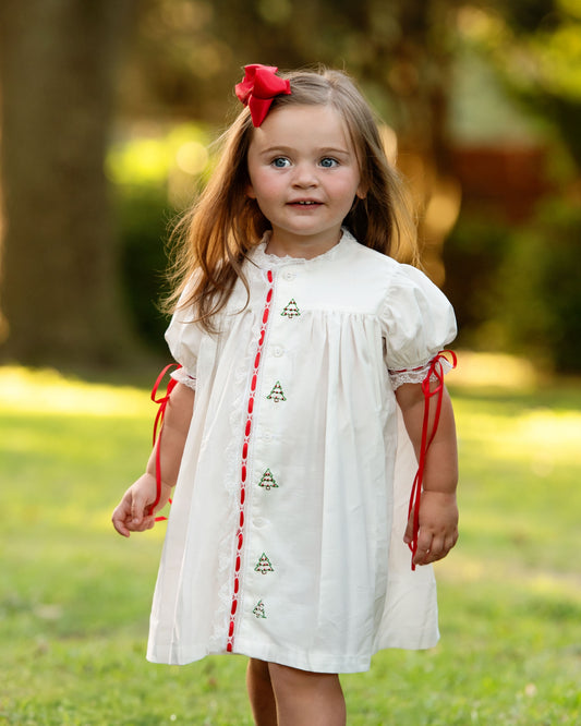A young girl wearing a white short-sleeve dress with red trim and tie details, standing outdoors on a grassy lawn with trees in the background.