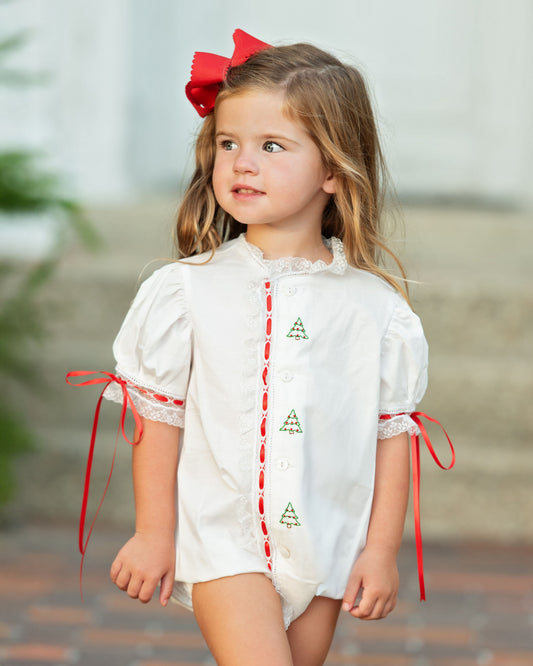 A young girl wearing a white short-sleeve romper with red trim and green embroidered Christmas tree motifs, accessorized with a red bow in her hair, set against a softly blurred outdoor background.