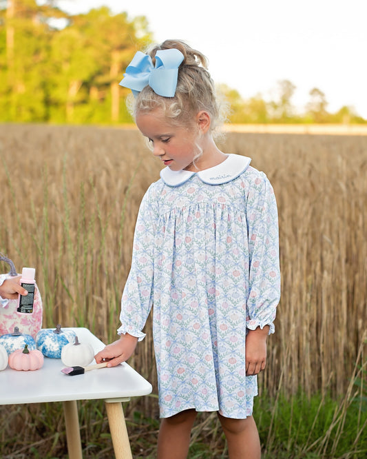 A young girl with a blonde ponytail and a large light blue bow stands outdoors in a wheat field, wearing a light blue and pink floral long-sleeve dress with a white Peter Pan collar. She is looking down at a small white table on which pumpkins and a phone are placed.