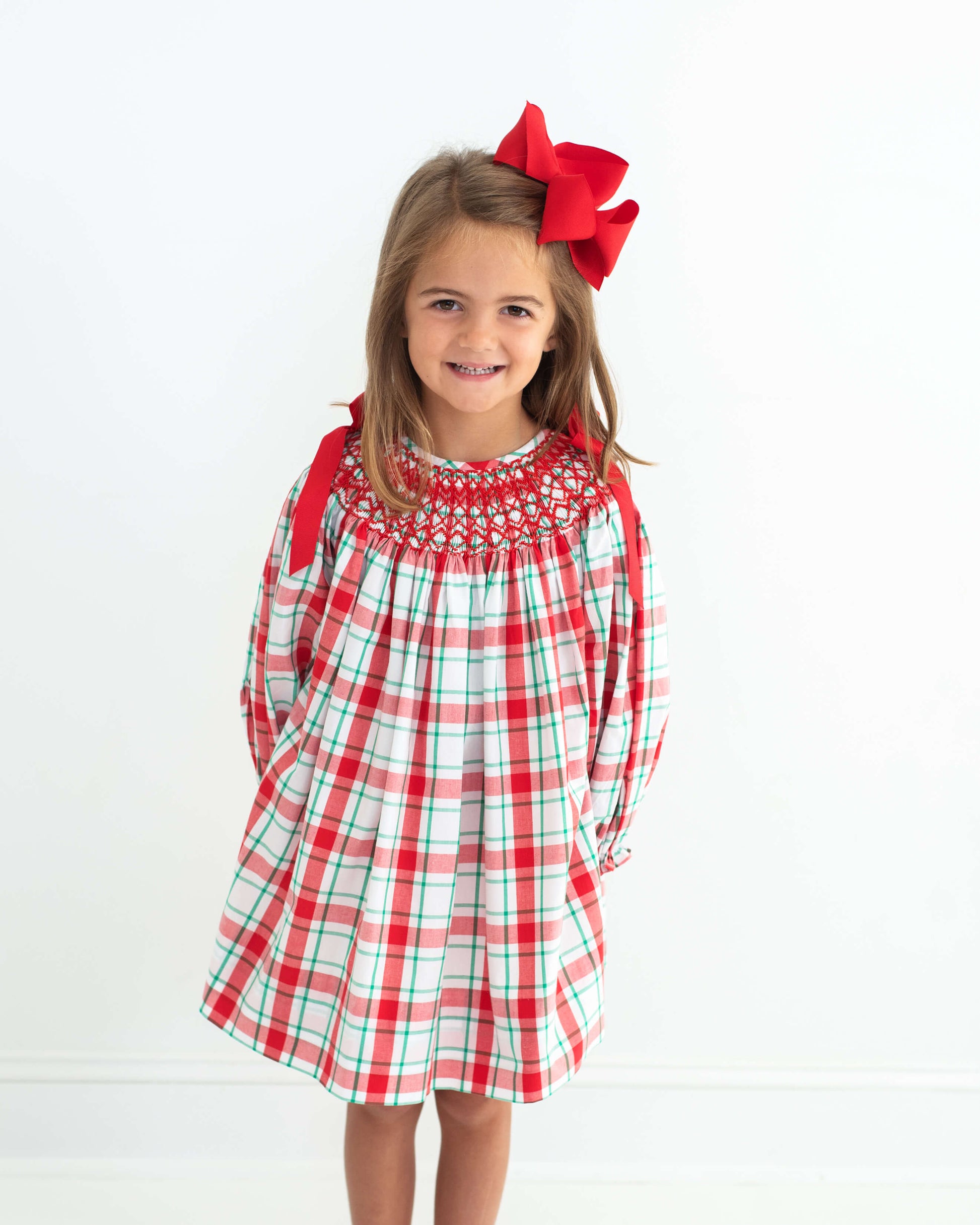 A young girl with a large red bow in her hair is wearing a red, white, and green plaid long-sleeve dress with smocked detailing on the chest, standing against a plain white background.