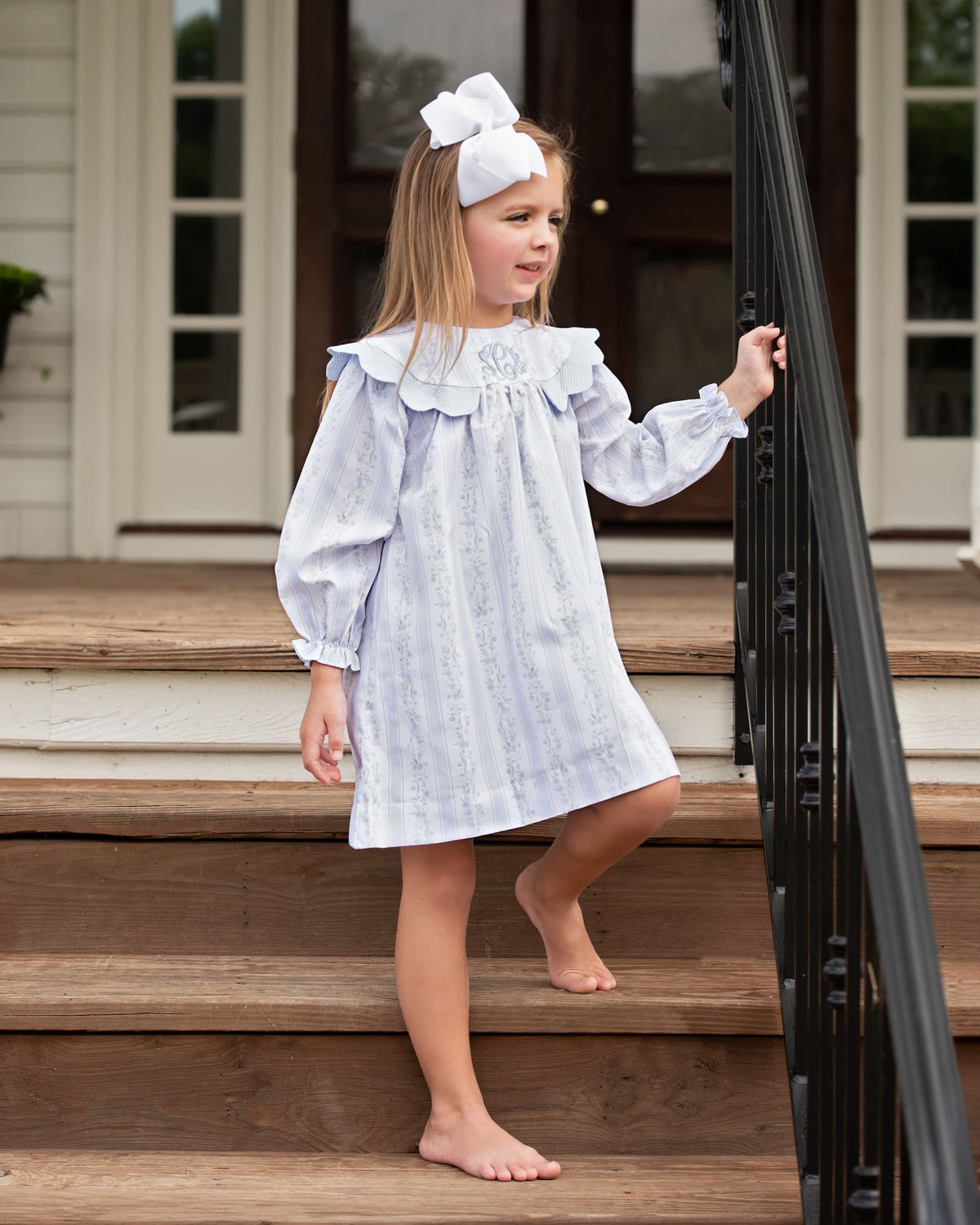A young girl with a large white bow in her hair stands barefoot on wooden steps next to a black metal railing, wearing a light blue and white patterned long-sleeve nightgown with a ruffled collar.