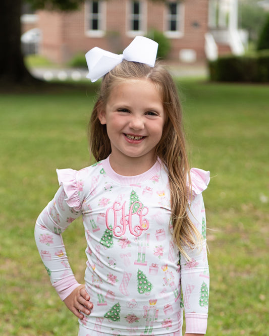 A young girl with a large white bow in her hair stands outdoors on a grassy lawn wearing a long-sleeve pajama top with a white base, pink ruffled shoulders, and a green and pink holiday print with a pink monogram on the chest.