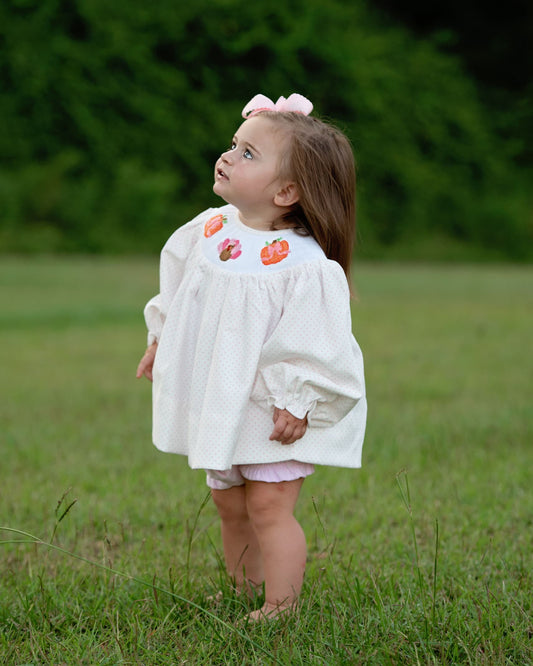 A young girl with a pink bow in her hair stands barefoot in a grassy field, wearing a white long-sleeve dress with colorful embroidered flowers on the chest.