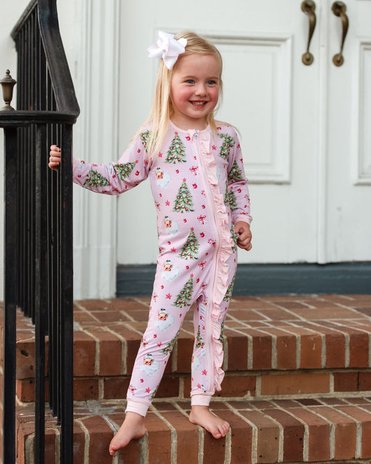 A young girl with a white bow in her hair stands barefoot on brick steps in front of a white door, wearing a light pink one-piece pajama with a green Christmas tree and red ornament print.