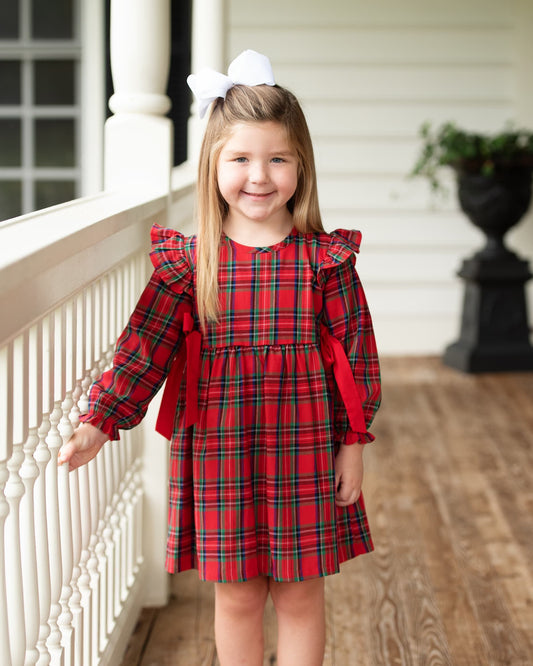 A young girl with a white bow in her hair stands on a porch wearing a red and green plaid dress with ruffled long sleeves and red side bows.