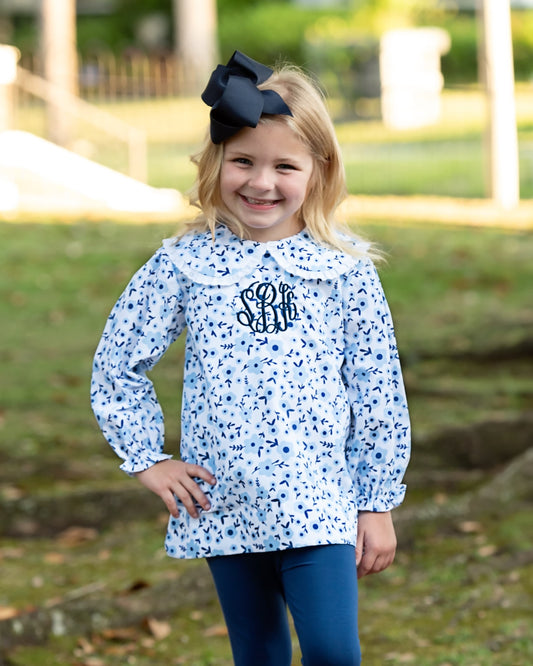 A young girl with blonde hair and a large black bow stands outdoors wearing a light blue long-sleeve blouse with a navy blue abstract print, a white Peter Pan collar, and a navy monogram on the chest. She is also wearing dark blue leggings.