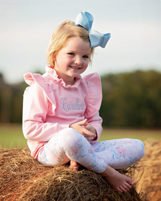 A young girl with blonde hair and a large light blue bow sits cross-legged on a bale of hay outdoors. She is wearing a pink long-sleeve ruffled top with the name 'Caroline' in blue script and white floral print leggings.