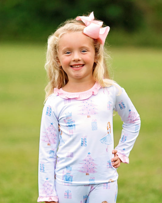 A young girl with blonde hair and a pink bow stands outdoors on a grassy lawn, wearing a light blue long-sleeve pajama top with a pink ruffled collar and a print of pink Christmas trees and blue presents, paired with matching light blue pajama pants.