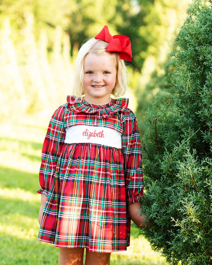 A young girl with blonde hair and a red bow stands outdoors next to a green tree, wearing a red plaid dress with a white chest panel embroidered with the name 'elizabeth' in red script.