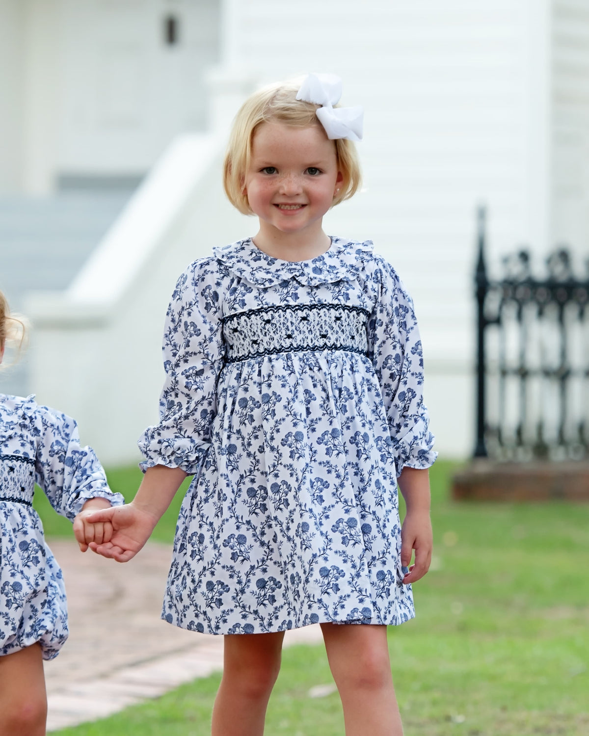 A young girl with blonde hair and a white bow stands outdoors holding hands with another child, wearing a blue and white floral smocked dress.