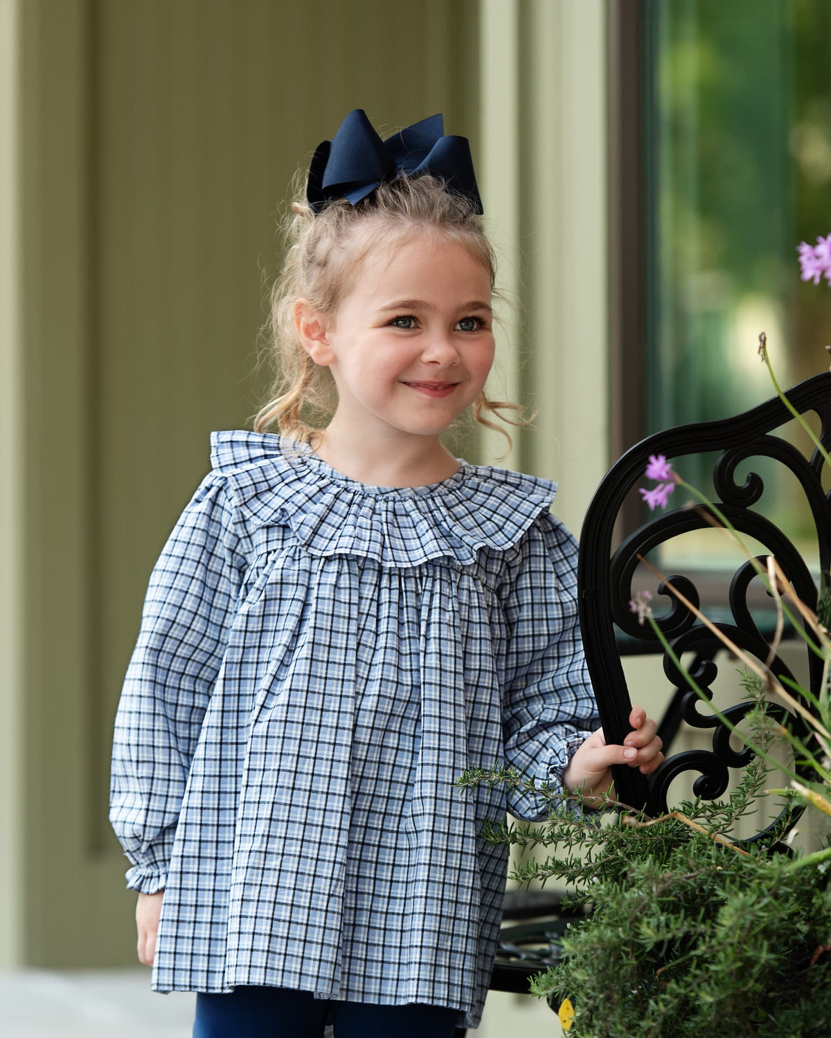 A young girl with blonde hair in a navy bow stands on a porch wearing a blue and white gingham long-sleeve top with a ruffled collar and dark blue pants.