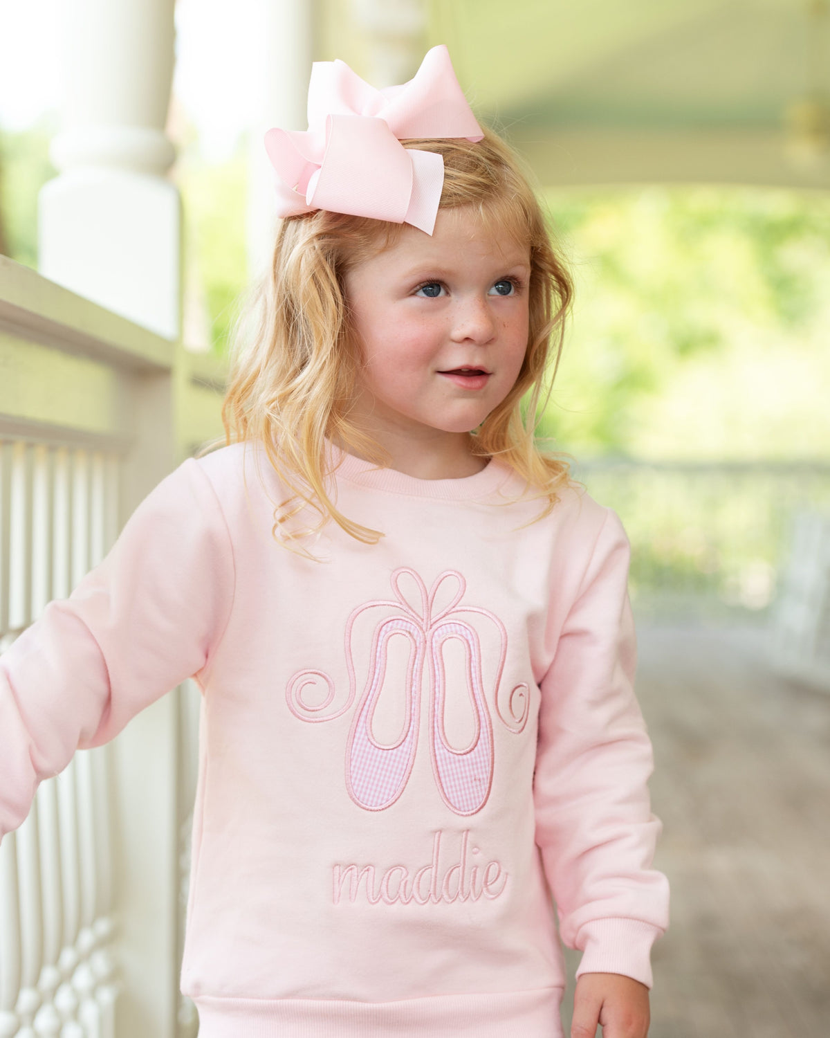 A young girl with blonde hair wearing a large light pink bow in her hair and a matching light pink sweatshirt with a ballet shoe graphic and the name 'maddie' embroidered on the front, standing on a porch.