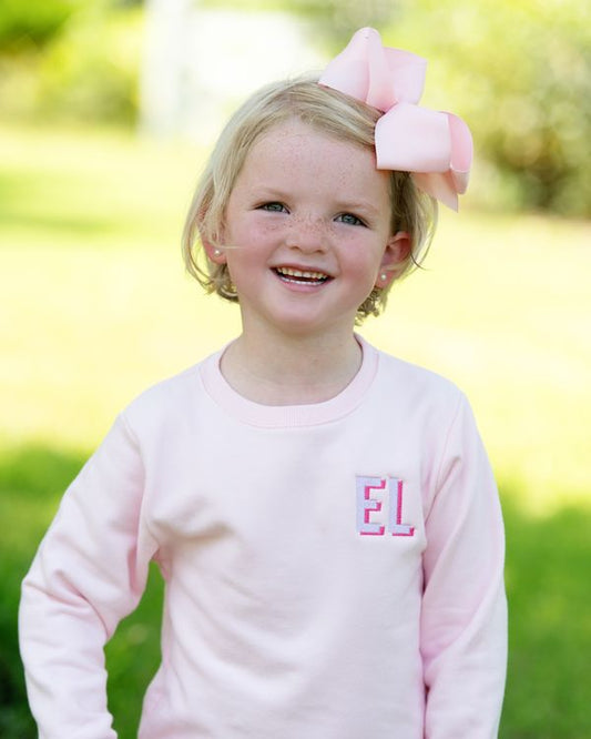 A young girl with blonde hair wearing a large pink bow in her hair and a light pink long-sleeve shirt with the letters 'EL' embroidered on the chest, standing outdoors in a grassy park setting.