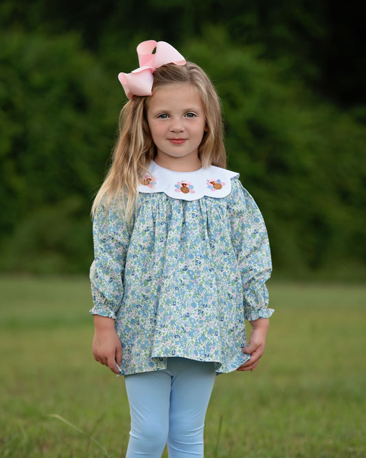 A young girl with blonde hair wearing a light blue and white floral long-sleeve blouse with a white Peter Pan collar featuring embroidered flowers, paired with light blue leggings and a pink bow, standing in a grassy outdoor setting.