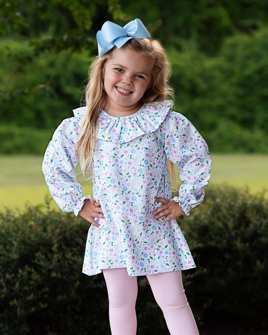 A young girl with blonde hair wearing a light blue bow, a white long-sleeve dress with a pink and green floral print, and light pink leggings, standing outdoors in front of a green hedge.