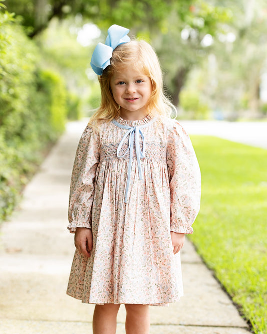 A young girl with blonde hair wearing a light blue bow stands on a paved path in a park, wearing a long-sleeve, knee-length dress with a delicate pink and blue floral pattern and a light blue ruffled collar with a tie.