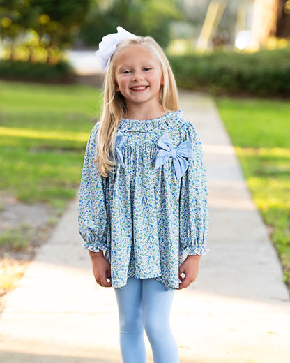A young girl with blonde hair wearing a light blue floral long-sleeve blouse with large bows on the chest, light blue leggings, and a white hair bow, standing on a concrete path in a sunny outdoor setting.