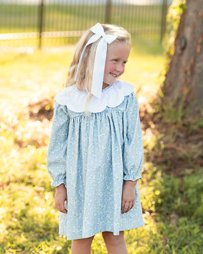 A young girl with blonde hair wearing a light blue patterned dress with a large white ruffled collar and a white hair bow, standing outdoors in a grassy yard.