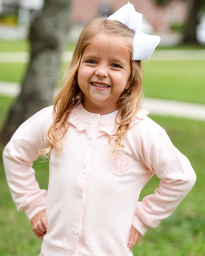 A young girl with blonde hair wearing a light pink long-sleeve button-up cardigan with a ruffled collar and a white bow, standing outdoors on a grassy lawn.