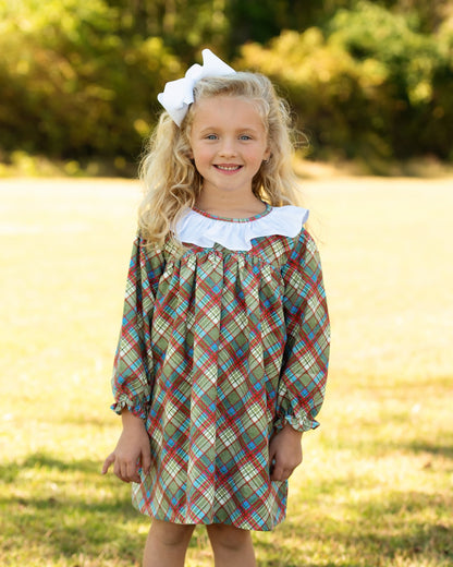 A young girl with blonde hair wearing a long-sleeve plaid dress with a white ruffled collar and a white bow, standing outdoors in a grassy field.