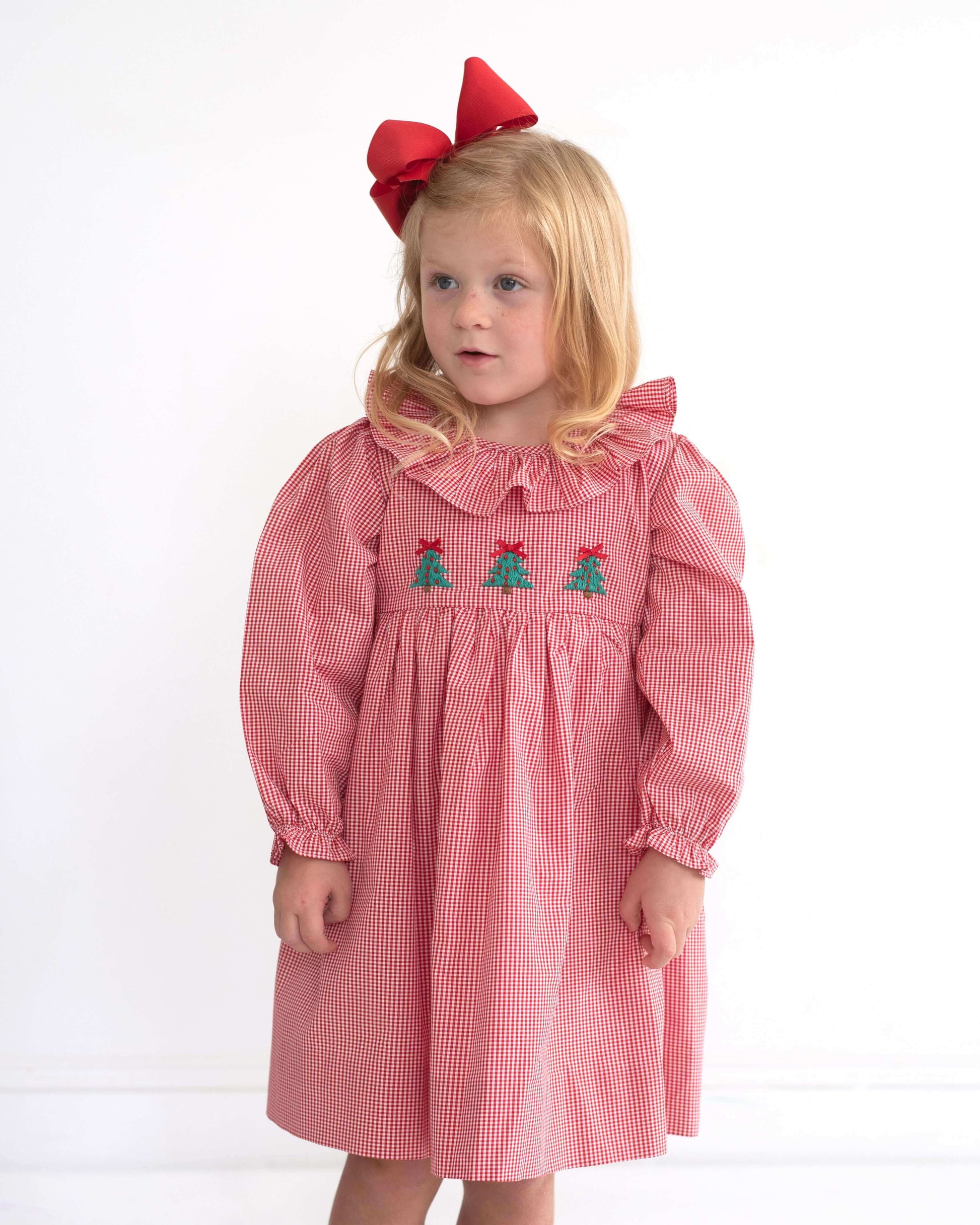 A young girl with blonde hair wearing a pink and white gingham dress with embroidered green trees and red bows on the chest, paired with a large red bow headband, standing against a plain white background.