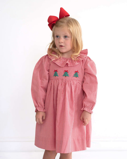 A young girl with blonde hair wearing a pink and white gingham dress with embroidered green trees and red bows on the chest, paired with a large red bow headband, standing against a plain white background.