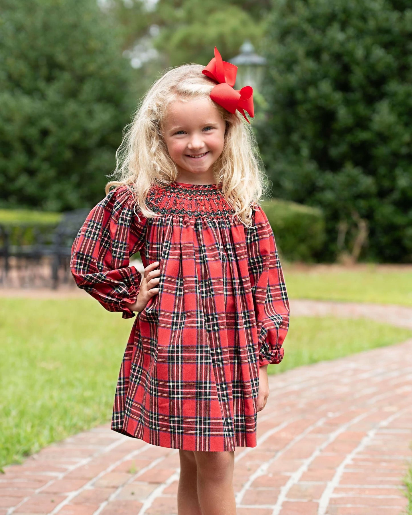 A young girl with blonde hair wearing a red and black plaid dress with a smocked bodice, standing on a brick path in a park with green trees in the background.