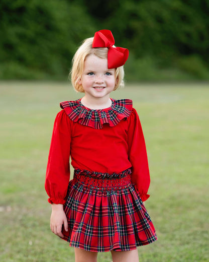 A young girl with blonde hair wearing a red long-sleeve top and matching red plaid skirt, accessorized with a large red bow, standing outdoors on a grassy lawn.