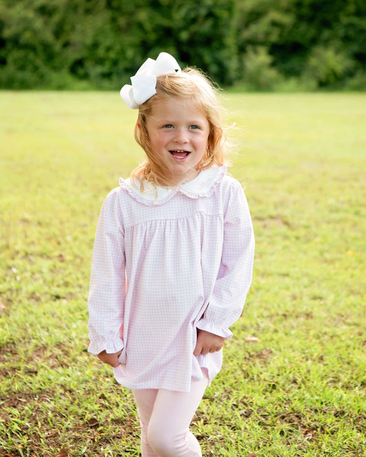 A young girl with blonde hair wearing a white bow stands in a grassy field, wearing a light pink and white gingham long-sleeve dress with a white ruffled collar and white leggings.