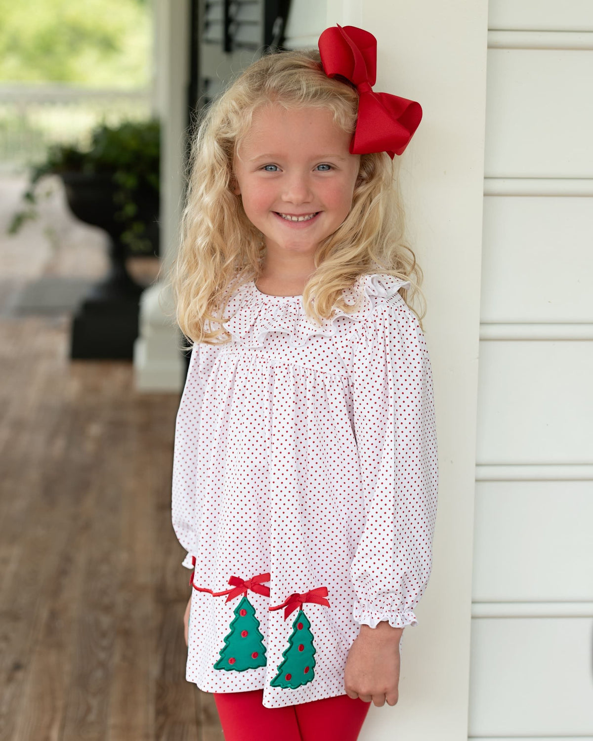 A young girl with blonde hair wearing a white long-sleeve blouse with red polka dots, red tights, and a large red bow in her hair, standing on a porch.