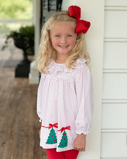 A young girl with blonde hair wearing a white long-sleeve blouse with red polka dots, red tights, and a large red bow in her hair, standing on a porch.
