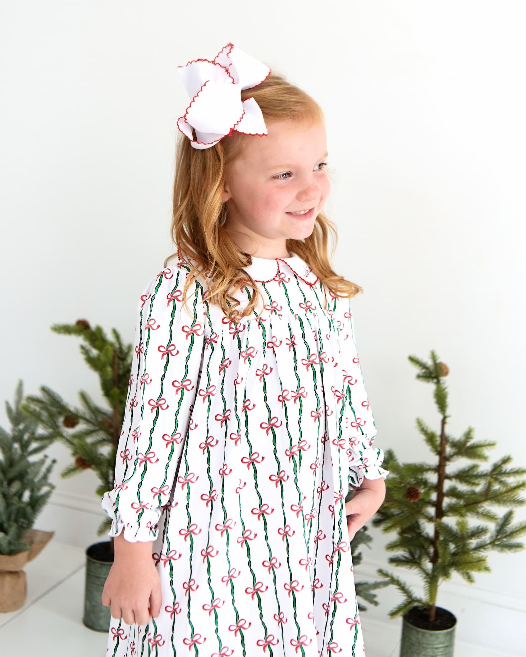 A young girl with blonde hair wearing a white long-sleeve dress with a green and red striped bow pattern, standing in front of a white wall with potted plants.
