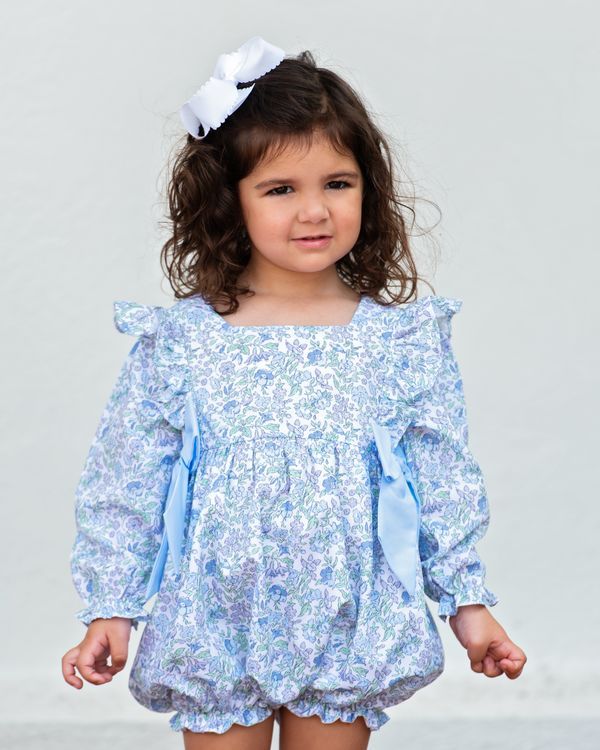 A young girl with curly brown hair wearing a light blue and white floral print long-sleeve romper with ruffled shoulders and a matching white bow headband, photographed against a plain light gray background.