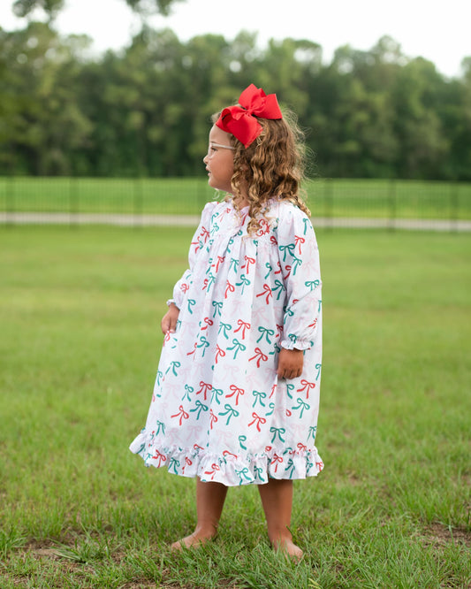A young girl with curly brown hair wearing a white long-sleeve dress with a red and green bow print, standing barefoot on a green grassy field. She is wearing a large red bow headband and looking to the side.