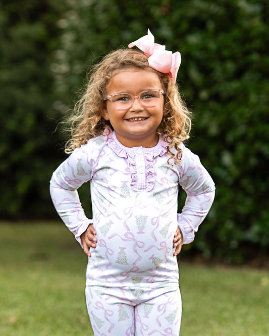 A young girl with curly hair, glasses, and a pink bow stands outdoors on grass wearing a light lavender long-sleeve pajama set with a pink and green Christmas tree pattern.