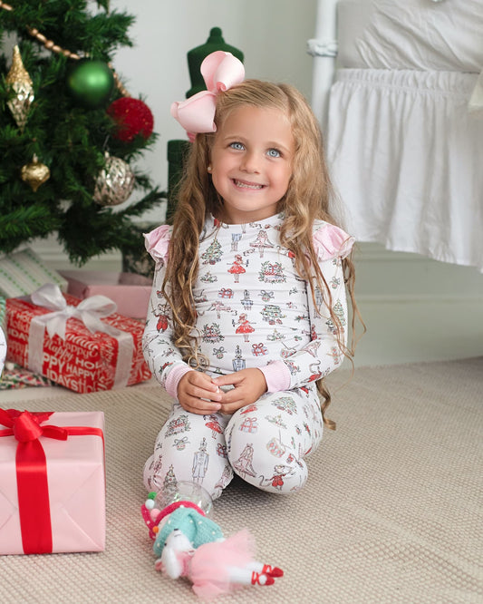 A young girl with long blonde hair sits on a carpeted floor in a cozy room decorated for Christmas. She is wearing a white long-sleeve pajama set with pink ruffled shoulders and a festive holiday print featuring Santa Claus, reindeer, and snowflakes.