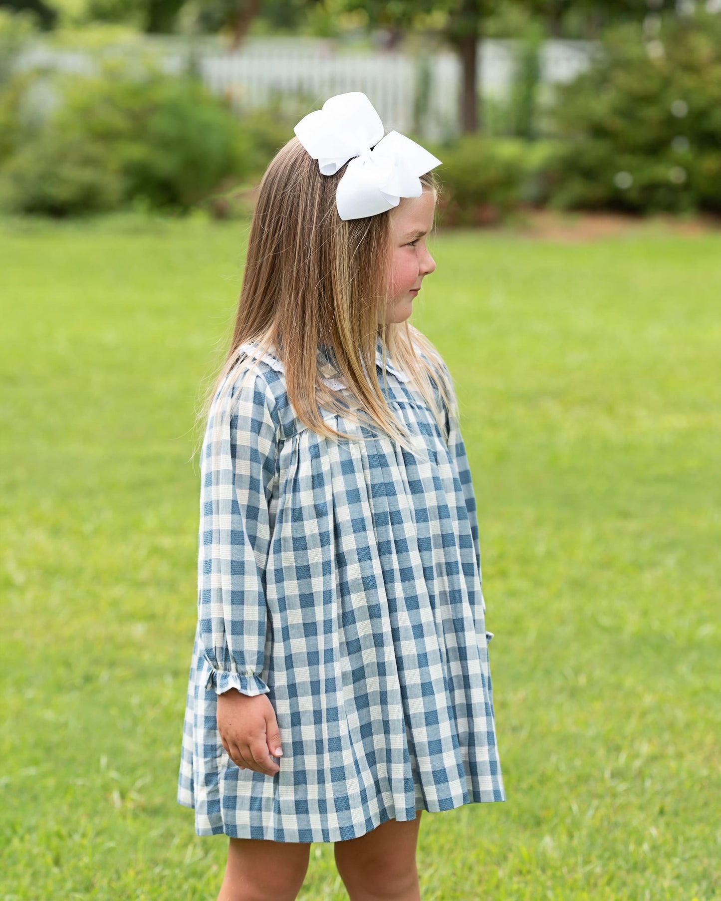 A young girl with long light brown hair wearing a blue and white gingham dress and a large white bow headband, standing on a green grassy lawn.