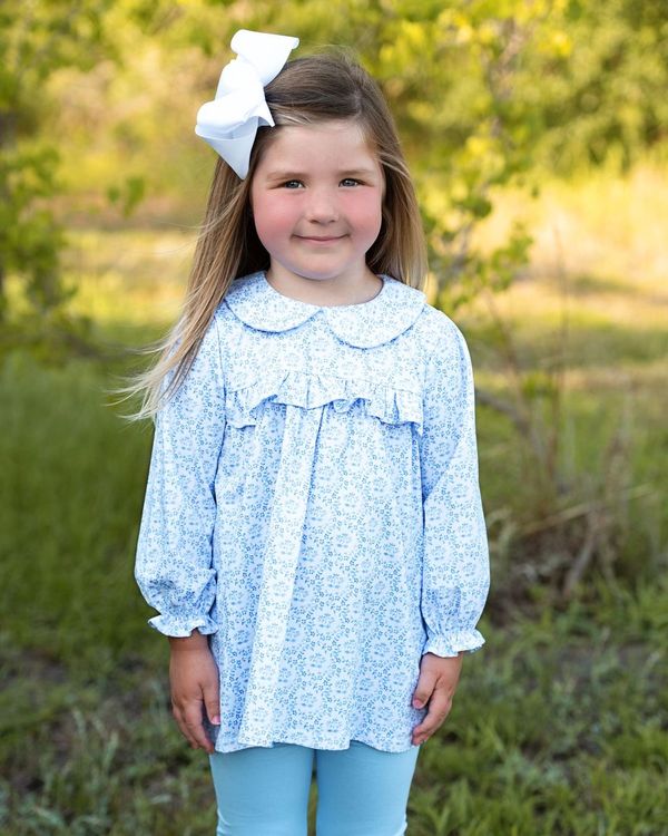A young girl with long light brown hair wearing a light blue and white floral long-sleeve top with a ruffled collar, a large white bow in her hair, and light blue leggings, standing outdoors in a grassy, wooded area.