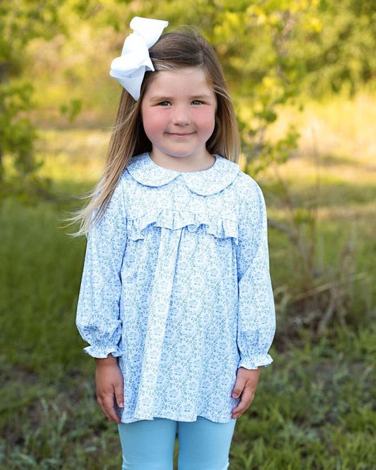 A young girl with long light brown hair wearing a light blue and white floral long-sleeve top with a ruffled collar, a large white bow in her hair, and light blue leggings, standing outdoors in a grassy, wooded area.