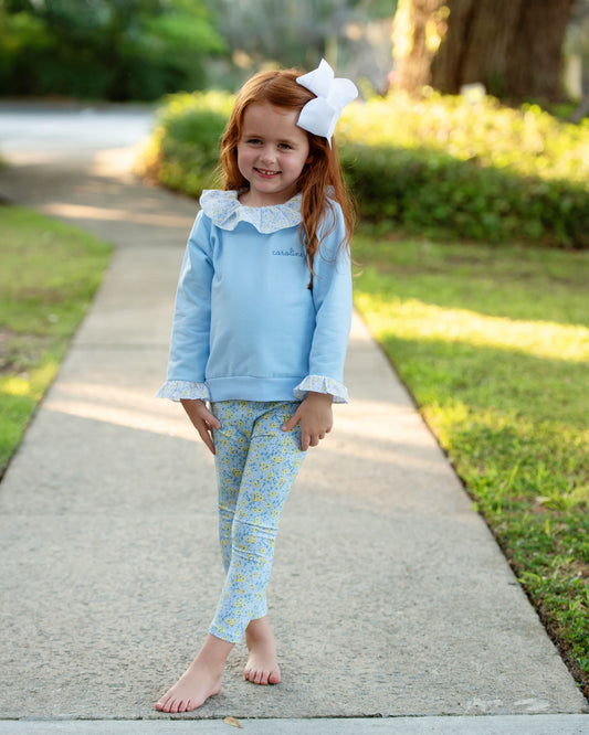 A young girl with red hair and a large white bow stands on a concrete path outdoors, wearing a light blue long-sleeve top with a white ruffled collar and matching blue patterned leggings.