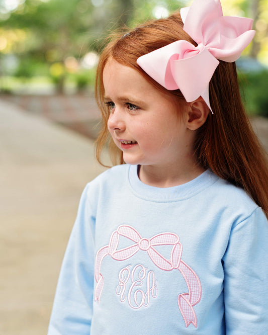 A young girl with red hair wearing a large pink bow in her hair and a light blue long-sleeve shirt with a pink ribbon and monogram design on the front, photographed outdoors on a blurred path.