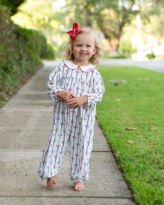 A young toddler girl stands barefoot on a concrete sidewalk outdoors, wearing a white long-sleeve footless jumpsuit with a red and black anchor pattern and a white Peter Pan collar. She has a red bow in her hair.