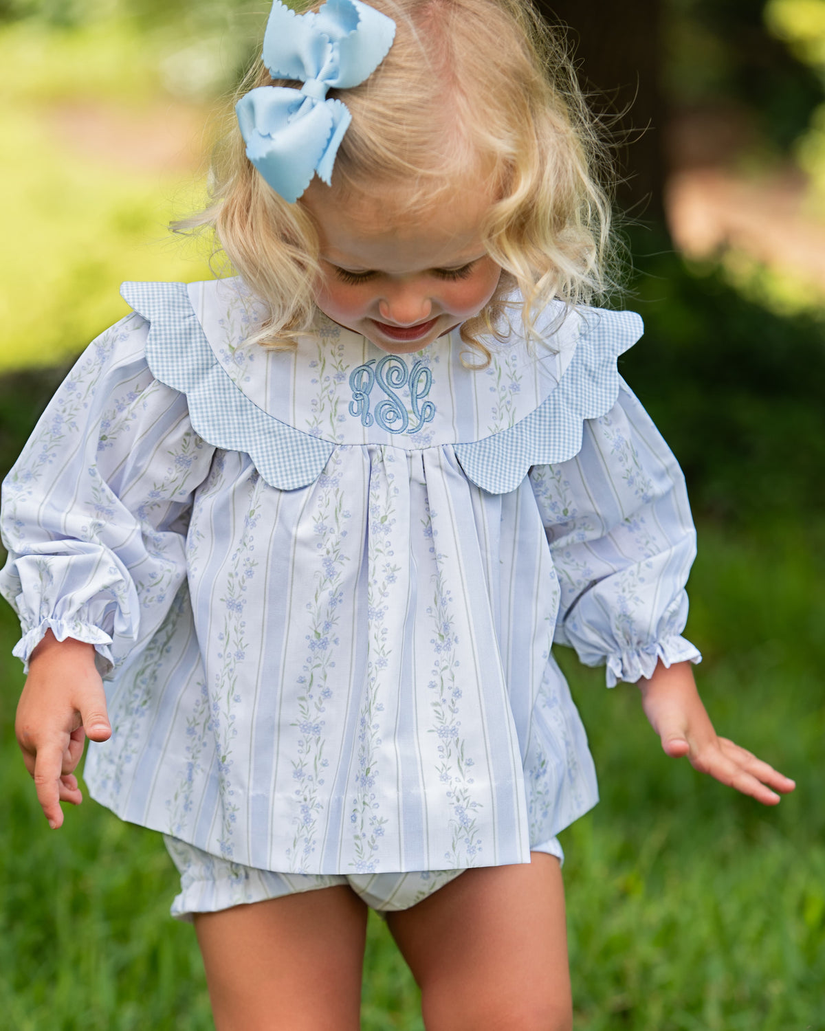 A young toddler girl wearing a light blue and white striped long-sleeve blouse with a ruffled collar and matching diaper cover, standing outdoors on a grassy lawn.